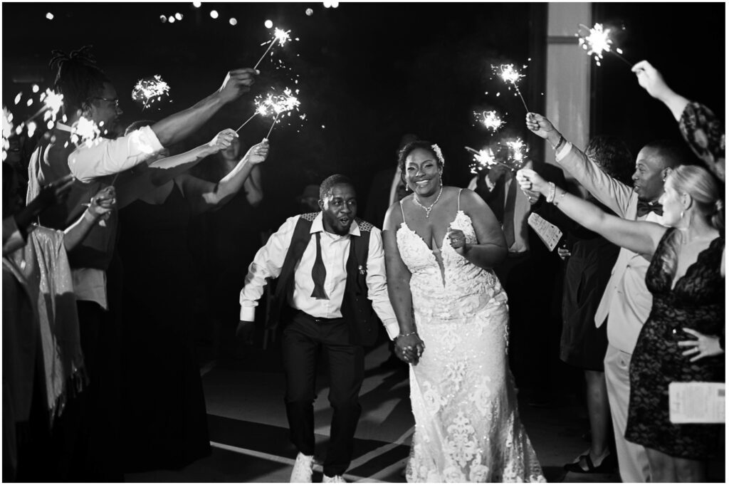A black and white photo of the bride and groom smiling while their guests hold sparklers around them. 